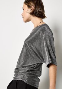 Silberner metallischer Kurzarm-Top mit lockerem Schnitt und Raffung in der Taille. Glatte Textur mit einem subtilen Schimmereffekt.