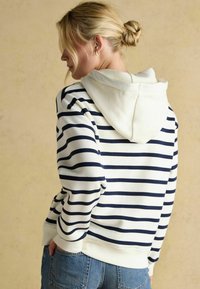 Kobieta z blond włosami upiętymi w kok, ubrana w białą bluzę z czarnymi poziomymi pasekami i niebieskie dżinsy, stojąca profil do beżowego tła.