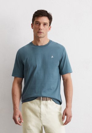 Blue cotton t-shirt with short sleeves, round neckline, and a small white logo on the chest, worn over beige linen trousers.