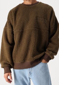 Brązowy polarowy sweatshirt o teksturowanej powierzchni, z opadającymi ramionami i brązowym ściągaczem na dole. Posiada subtelne logo na klatce piersiowej.