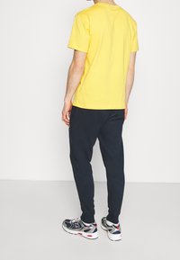 Geel katoenen T-shirt met korte mouwen, gecombineerd met marineblauwe joggers. De persoon draagt grijze en rode sportschoenen met een gaasstructuur.