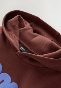 Gros plan sur un sweat à capuche marron avec une capuche rembourrée et un texte bleu partiellement visible imprimé sur le tissu.