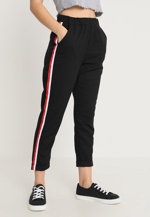 Persoon draagt zwarte joggingbroek met rode en witte zijstrepen, gecombineerd met zwarte sneakers en een kort grijs topje.