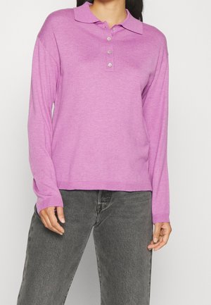 Femme portant un polo à manches longues violet clair et un jean gris foncé, debout devant un fond uni.