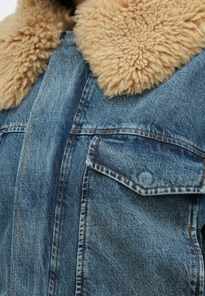 Džinsinė striukė su dideliu rudu avies kailio apykakle ir priekinė kišenė su atvartais, rodanti detalią tekstūrą ir siuvimo linijas.