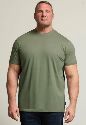 Homme aux cheveux courts portant un t-shirt vert olive à manches courtes et un pantalon foncé, debout devant un fond beige uni.