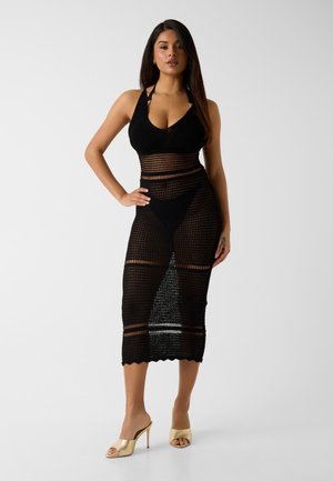 Kobieta stoi, ubrana w czarną szydełkowaną sukienkę na szyję, na czarnej bieliźnie i złote, wysokie, odkryte mules na białym tle.