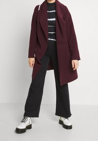 Abrigo burdeos de corte abierto con diseño drapeado y mangas largas, llevado sobre una camiseta negra a rayas, acompañado de pantalones anchos negros y botas blancas.