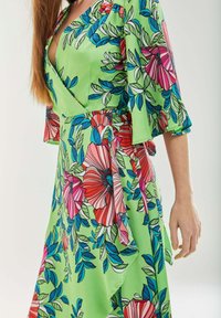 Floral wrap dress in green fabric, featuring pink, red, and blue flowers with leaf patterns. Highlighted by flared sleeves and a flowing silhouette.