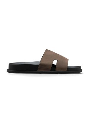Sandale mules en cuir marron avec une semelle plate noire et une large bride présentant une découpe rectangulaire près de la voûte plantaire.