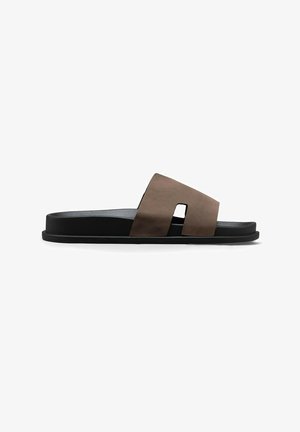 Sandale mules en cuir marron avec une semelle plate noire et une large bride présentant une découpe rectangulaire près de la voûte plantaire.