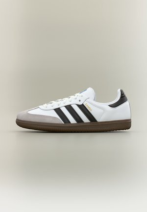 Baskets Adidas Samba blanches avec des bandes noires, bout en daim gris et semelle marron, vues de côté sur un fond neutre.