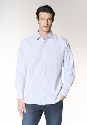 Uomo che indossa una camicia a righe azzurro chiaro e bianco a maniche lunghe con bottoni e jeans blu scuro, in piedi con una mano in tasca.