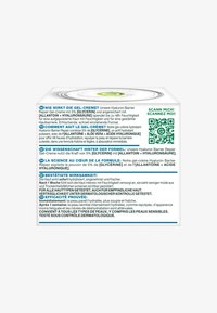 Biały kwadratowy pojemnik z zielonymi akcentami, zawierający informacje o produkcie w wielu językach, kod QR oraz graficzny okrągły design pokrywy.