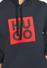Sudadera navy con cordones negros, que cuenta con un cuadrado rojo con grandes letras negras "HUGO" en el pecho y un bolsillo delantero.
