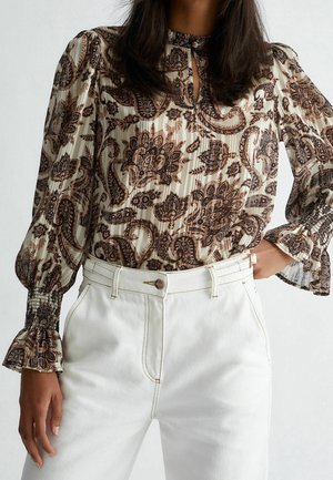 Femme portant un chemisier marron à manches longues à motifs avec poignets smockés et un pantalon blanc taille haute, main sur la hanche.