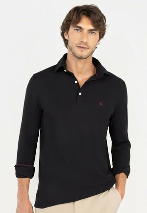 Joven con cabello castaño claro, vistiendo una camisa polo negra de manga larga y pantalones beige, de pie con una mano en el bolsillo.