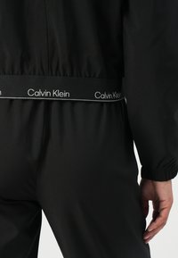 Zwarte trainingsbroek met elastische tailleband met een witte "Calvin Klein" logo. Gladde textuur, comfortabele pasvorm met verknoping bij de manchetten.