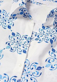 Camisa de tela blanca con intrincados bordados florales azules, con un cuello puntiagudo y un solo botón de perla en la parte delantera.