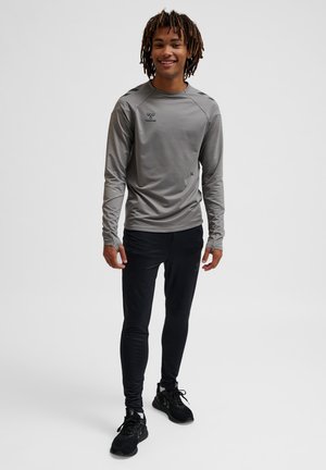 Maglia sportiva a maniche lunghe grigia con logo sul petto, abbinata a leggings neri aderenti e scarpe sportive nere, in piedi su uno sfondo neutro.