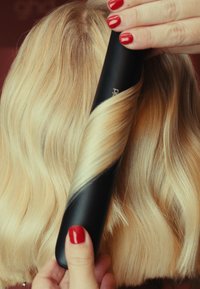 Schwarzer Lockenstab vor langem, welligem blondem Haar positioniert, der ein glattes Finish und eine glänzende Textur zeigt. Rote, manikürte Nägel halten den Lockenstab.