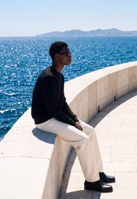 Homme en pull noir, pantalon blanc et lunettes de soleil assis sur un banc en pierre courbé au bord de la mer bleue étincelante sous un ciel clair.