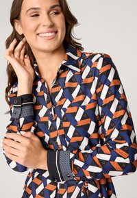 Overhemd met knoopsluiting, voorzien van een geometrisch patroon in blauw, oranje en wit. Zwarte manchetten met een decoratieve afwerking, zachte stoftextuur.