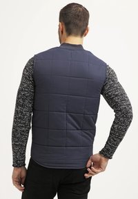 Marinblå quiltad väst, utan ärmar, med en enkel design; bärs över en långärmad spräcklig svart och vit skjorta. Blandning av bomull.