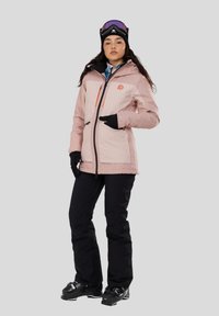 Lys pink ski-jakke med sorte detaljer og lynlås, parret med sorte skibukser og støvler. Modellen bærer en sort hue og skibriller.