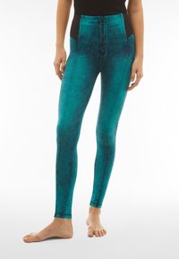 Leggings color teal con un alto cinturino nero, caratterizzati da una zip frontale e una finitura testurizzata, progettati per una silhouette aderente e attillata.