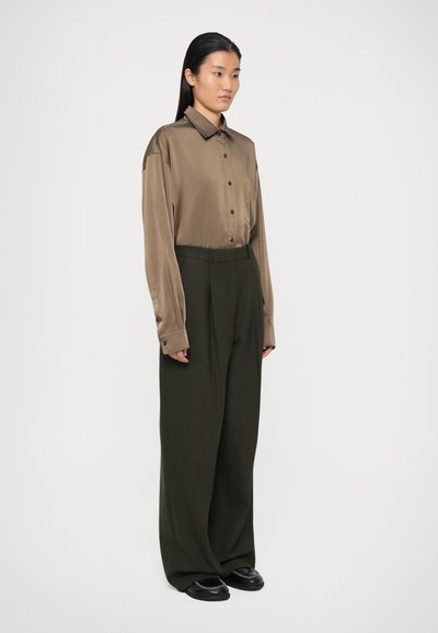 Cămașă maro, cu mânecă lungă și nasturi, având un buzunar; asortată cu pantaloni largi, de culoare verde închis; modelul poartă pantofi negri cu accente decorative.