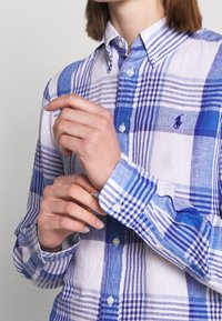 Camicia a quadri blu e bianca con bottoni in tessuto leggero. Presenta quadri a contrasto, un logo sul petto e polsini con bottoni.