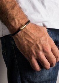 Black braided leather bracelet with a gold cylindrical clasp, worn on a wrist, featuring a textured finish and contrasting colors.