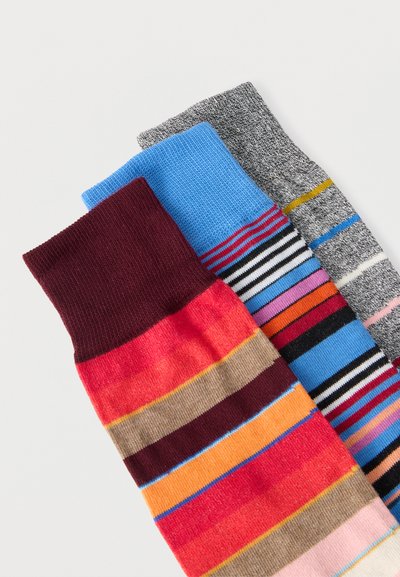 Trois paires de chaussettes rayées colorées dans des tons bordeaux, bleu et gris, disposées en chevauchement sur un fond blanc.