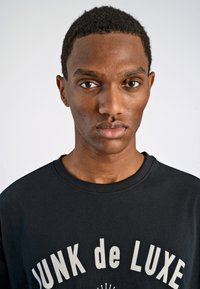 Camiseta de algodón negra con "JUNK de LUXE" impreso en color crema, diseño de cuello redondo, mangas cortas y un ajuste relajado. Textura suave.