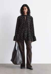 Schwarze Bluse mit floralen Mustern, langen Ärmeln und Schlüsselloch-Ausschnitt, kombiniert mit dunkelbraunen, weiten Hosen und einer großen grauen Leder-Tasche.