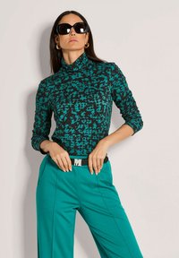 Türaquoisefarbener und schwarzer gemusterter Rollkragenpullover, kombiniert mit türkisfarbenen Hosen und einem schwarzen Gürtel mit einer silbernen Schnalle, vor einem neutralen Hintergrund präsentiert.