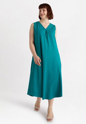 Türkises ärmelloses Kleid mit V-Ausschnitt, das eine dezente Vorderfalte und eine fließende Silhouette aufweist, kombiniert mit weißen Sandalen mit Riemchen.