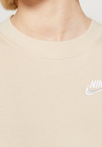 Beige sweatshirt med rund halsringning och en liten vit Nike-logotyp. Mjuk textur med en ren, minimalistisk design.