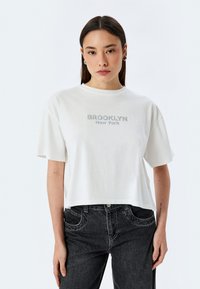 Camiseta blanca corta de algodón, con mangas cortas y cuello redondo. Texto gráfico "BROOKLYN New York" en gris en el pecho.