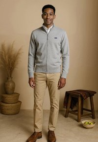 Grijze ritssweater met geribbelde manchetten, gedragen over een witte blouse met kraag, gecombineerd met khaki broek en bruine schoenen. Eenvoudig ontwerp en textuur.