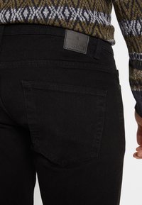 Gros plan d'une personne portant un jean noir et un pull en maille à motifs, montrant la poche arrière et une partie de la main droite.