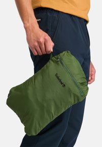 Groene, gevoerde tas van nylon met een ritssluiting en het Timberland-logo. Deze wordt vastgehouden door een persoon die een marineblauwe broek draagt.