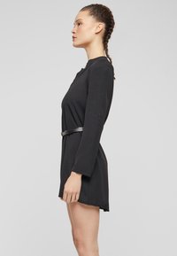 Zwarte jurk met lange mouwen, een ontspannen pasvorm en een riem in de taille. De stof heeft een gladde textuur, met een ronde halslijn en een zij-split.