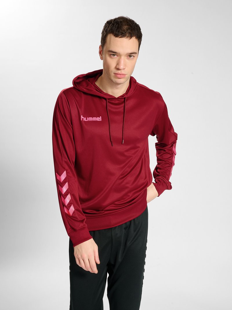 Junger Mann trägt einen roten Hoodie mit pinken Chevron-Streifen am Ärmel und schwarze Hose, steht mit einer Hand in der Tasche vor weißem Hintergrund.