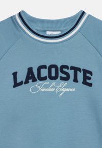 Sweat-shirt bleu avec col côtelé comportant des rayures blanches et marine et le texte "LACOSTE Timeless Elegance" brodé à l'avant.
