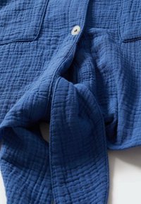 Sininen puuvillapaita, jossa on teksturoitu, tikattu kuvio, kaksi taskua ja yksittäinen nappikiinnitys. Pehmeä, rento kangas laskeutuu löysästi.