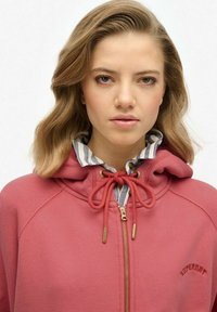 Sweat à capuche rouge avec fermeture éclair et finitions dorées, comportant un cordon de réglage à la capuche et un logo cousu visible. Superposé à une chemise rayée.