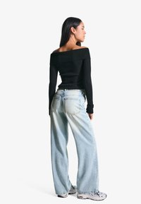 Haut noir à épaule dénudée en côtes associé à un jean évasé bleu clair. Le modèle porte des chaussures de sport argentées, mettant en avant un style décontracté.