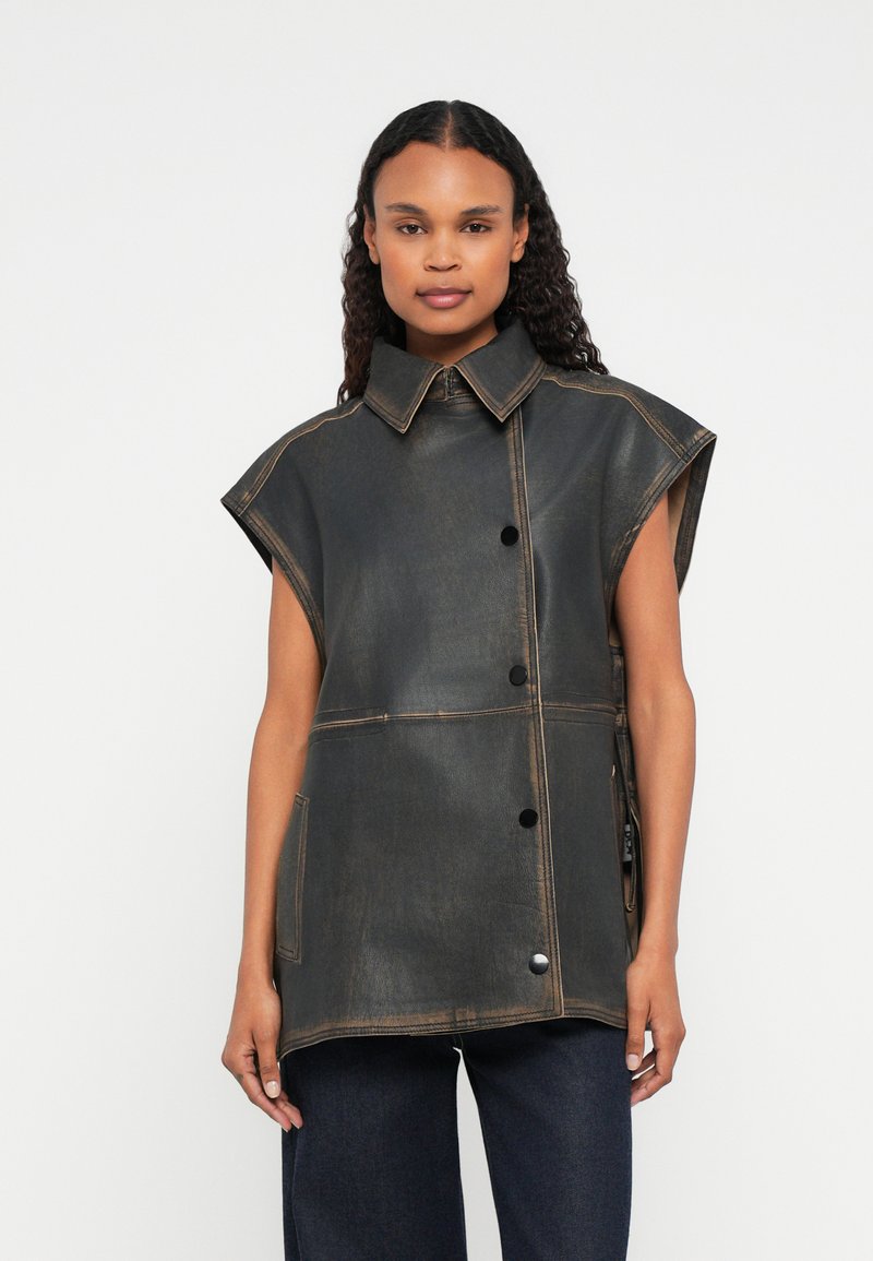 Donna che indossa un gilet in pelle marrone scuro senza maniche con bottoni a scatto e ampio collare, abbinato a pantaloni scuri su uno sfondo neutro.